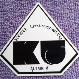 May include: A white diamond-shaped sticker with black text that reads "Krell University" and "KU" in a stylized font. The sticker also has the text "Altair V" in black.
