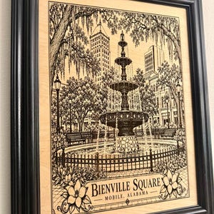 May include: A black-framed wooden wall art piece featuring a detailed black ink illustration of the Bienville Square fountain in Mobile, Alabama. The artwork depicts the tiered fountain, surrounding trees with Spanish moss, and city buildings.