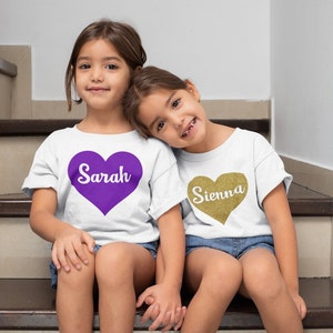 May include: Two young girls wearing white t-shirts with heart designs. One shirt has a purple heart with the name "Sarah" in white, and the other has a gold glitter heart with the name "Sienna" in white. They are sitting on wooden stairs.