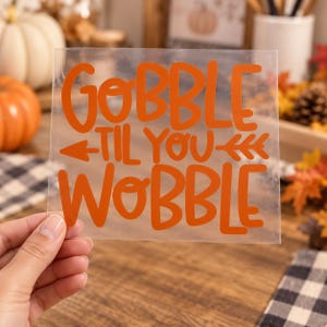 May include: A clear, rectangular adhesive decal featuring the orange text GOBBLE TIL YOU WOBBLE in a bold, rounded font. The design includes a horizontal arrow graphic pointing left and right, positioned between the words TIL YOU and WOBBLE.