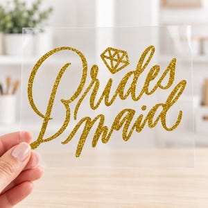 May include: A clear adhesive decal featuring the word Bridesmaid written in a gold glitter script font. Above the word is a small gold glitter diamond icon. The design is printed on a transparent backing sheet, ready for application to a surface.