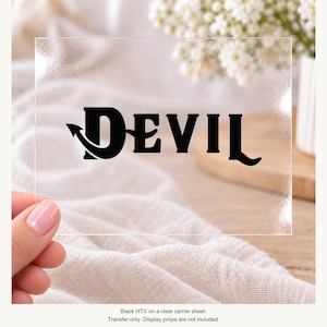 May include: A black heat transfer vinyl decal featuring the word DEVIL in a stylized font, where the letter D incorporates a pointed tail design. The black lettering is applied to a clear, rectangular carrier sheet for application purposes.