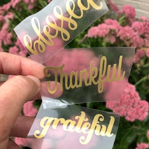 May include: Three individual clear adhesive decals featuring metallic gold lettering in a cursive script font. The top decal displays the word blessed, the middle decal shows the word Thankful, and the bottom decal features the word grateful.