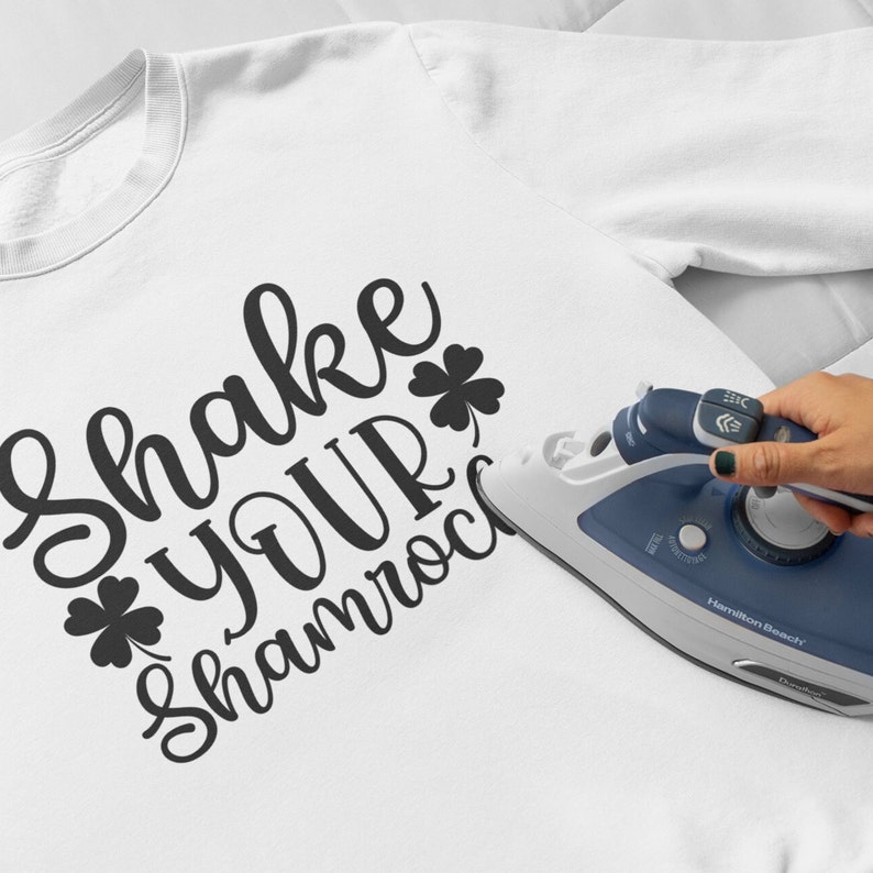 shake your shamrocks iron on decal shown in black being ironed on with a household iron onto a white tshirt