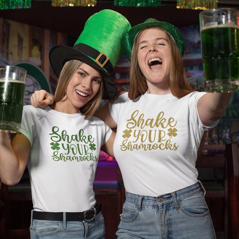 two women in a bar wearing shirts with shake your shamrocks text design holding green drinks and smiling