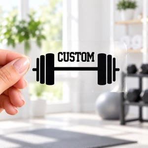 May include: A clear adhesive decal featuring a black graphic of a barbell with weight plates on each side. The word CUSTOM is printed in a bold, black, sans-serif font positioned directly above the center bar of the barbell graphic.