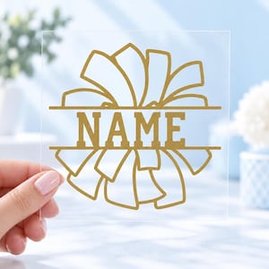 May include: A clear square decal featuring a gold-colored graphic of a cheerleading bow with a horizontal split in the center. The word NAME is printed in bold, gold, block capital letters within the split section of the bow design.