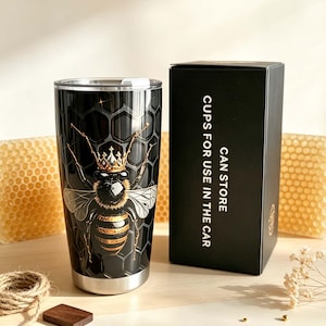 May include: A black stainless steel tumbler featuring a graphic of a bee wearing a gold crown against a honeycomb pattern. Next to it is a black rectangular box with the white text CAN STORE CUPS FOR USE IN THE CAR printed on the side.