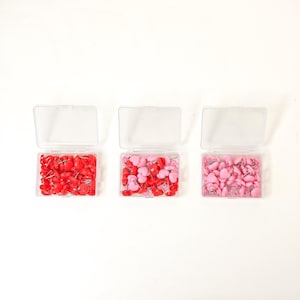 May include: Three clear plastic containers filled with heart-shaped push pins. The containers are arranged in a row from left to right. The push pins in the left container are red, the push pins in the middle container are pink, and the push pins in the right container are light pink.