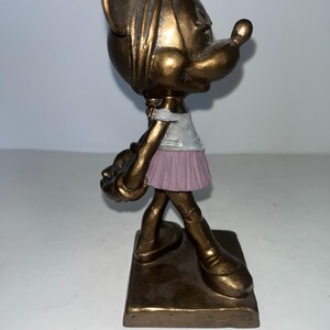 May include: A bronze-effect figurine of a cartoon character, wearing a white top and a pink skirt. The figurine stands on a rectangular base and holds a small object. The character has large ears and a prominent nose.