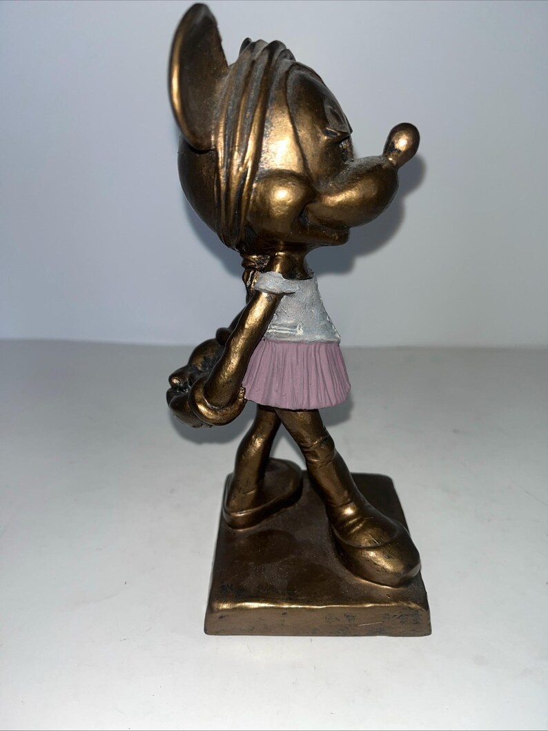 May include: A bronze-effect figurine of a cartoon character, wearing a white top and a pink skirt. The figurine stands on a rectangular base and holds a small object. The character has large ears and a prominent nose.