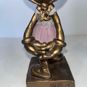 May include: A bronze-coloured figurine of a character wearing a pink dress and a white scarf. The figurine has large, rounded hands and feet, and the base is rectangular. The Disney logo is visible on the base.