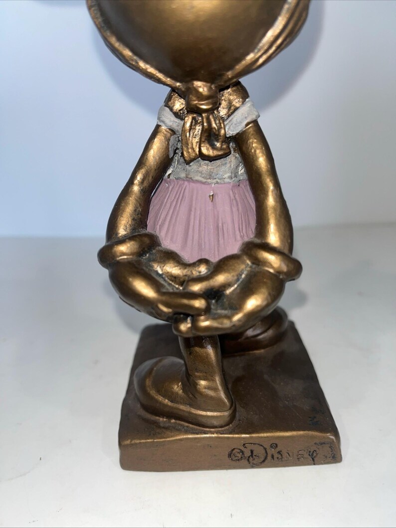 May include: A bronze-coloured figurine of a character wearing a pink dress and a white scarf. The figurine has large, rounded hands and feet, and the base is rectangular. The Disney logo is visible on the base.