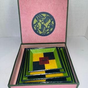 Könnte beinhalten: Ein Satz von vier quadratischen dekorativen Tabletts, die in einer rosa Box mit grüner Umrandung verschachtelt sind. Die Tabletts haben ein grün-gelb-schwarzes Schachbrettmuster mit einem roten Symbol in der Mitte. Der Deckel der Box hat ein kreisförmiges Design.