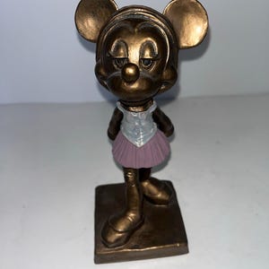 May include: A bronze-coloured figurine of Minnie Mouse in a ballet outfit. The figurine has a square base and features Minnie's iconic ears, a light purple tutu, and a white top.