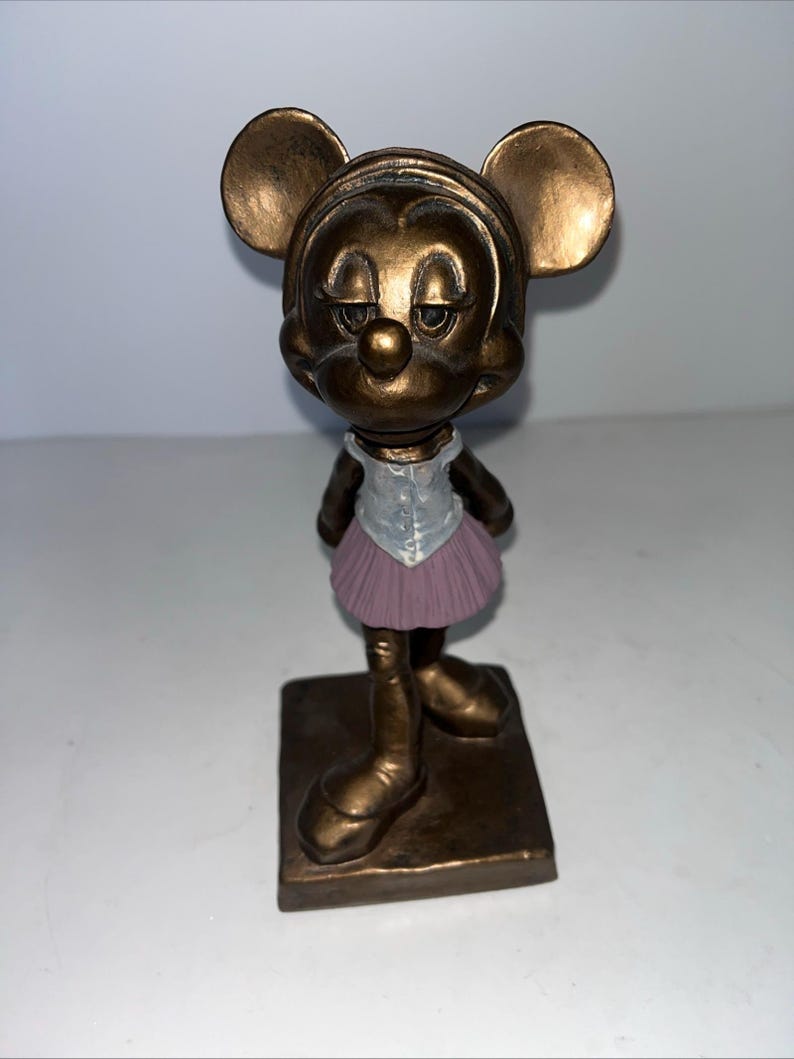 May include: A bronze-coloured figurine of Minnie Mouse in a ballet outfit. The figurine has a square base and features Minnie's iconic ears, a light purple tutu, and a white top.