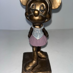 May include: A bronze-effect figurine of a mouse character in a ballet outfit. The figurine has large ears, a white top, a pink tutu, and is standing on a square base. The mouse has a slightly sad expression.