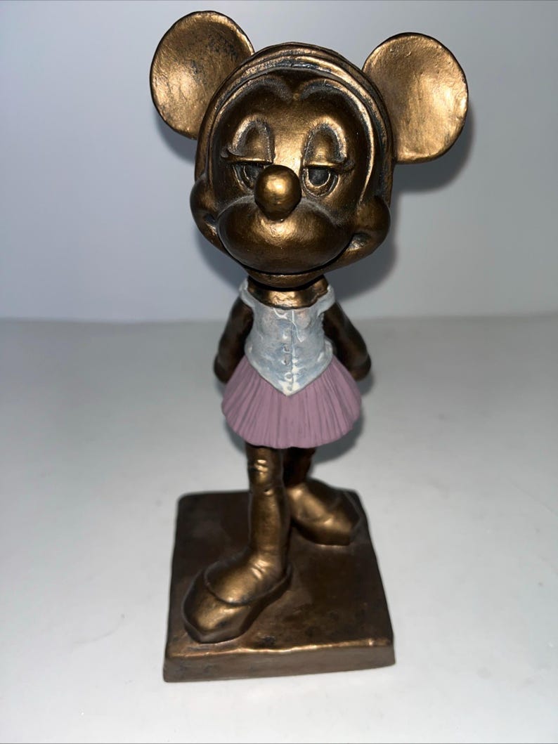 May include: A bronze-effect figurine of a mouse character in a ballet outfit. The figurine has large ears, a white top, a pink tutu, and is standing on a square base. The mouse has a slightly sad expression.
