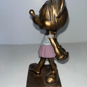 May include: A bronze-coloured figurine of a cartoon character, wearing a light blue top and a pink skirt. The figure is standing on a rectangular base. The figurine has large ears and a long nose.