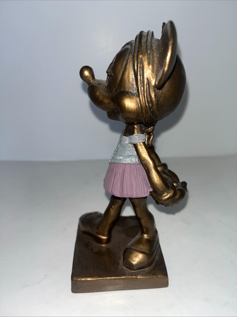 May include: A bronze-coloured figurine of a cartoon character, wearing a light blue top and a pink skirt. The figure is standing on a rectangular base. The figurine has large ears and a long nose.