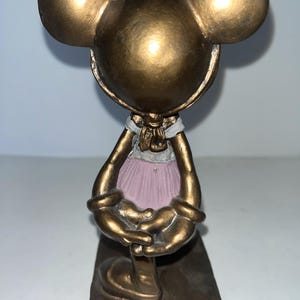 May include: A bronze-coloured Mickey Mouse figurine in a ballet outfit. The figurine has large round ears, a white collar, and a pink tutu. The base of the figurine is rectangular and has the Disney logo.