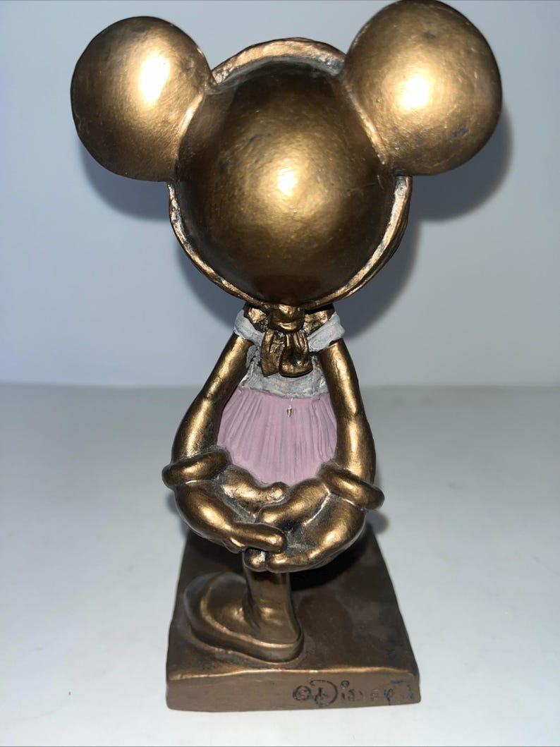 May include: A bronze-coloured Mickey Mouse figurine in a ballet outfit. The figurine has large round ears, a white collar, and a pink tutu. The base of the figurine is rectangular and has the Disney logo.