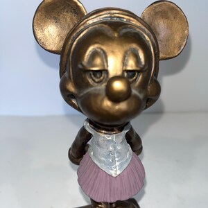 May include: A bronze-coloured figurine of a mouse character wearing a light blue vest and a pink tutu. The mouse has large, round ears and a slightly sad expression. The figurine is standing on a rectangular base.