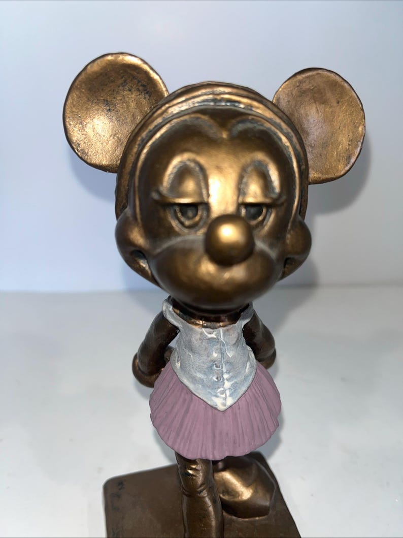 May include: A bronze-coloured figurine of a mouse character wearing a light blue vest and a pink tutu. The mouse has large, round ears and a slightly sad expression. The figurine is standing on a rectangular base.