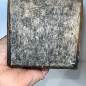 May include: Close-up of a weathered, rectangular wooden block with a dark, textured surface. The edges are framed with a darker wood. The block is held in a hand.