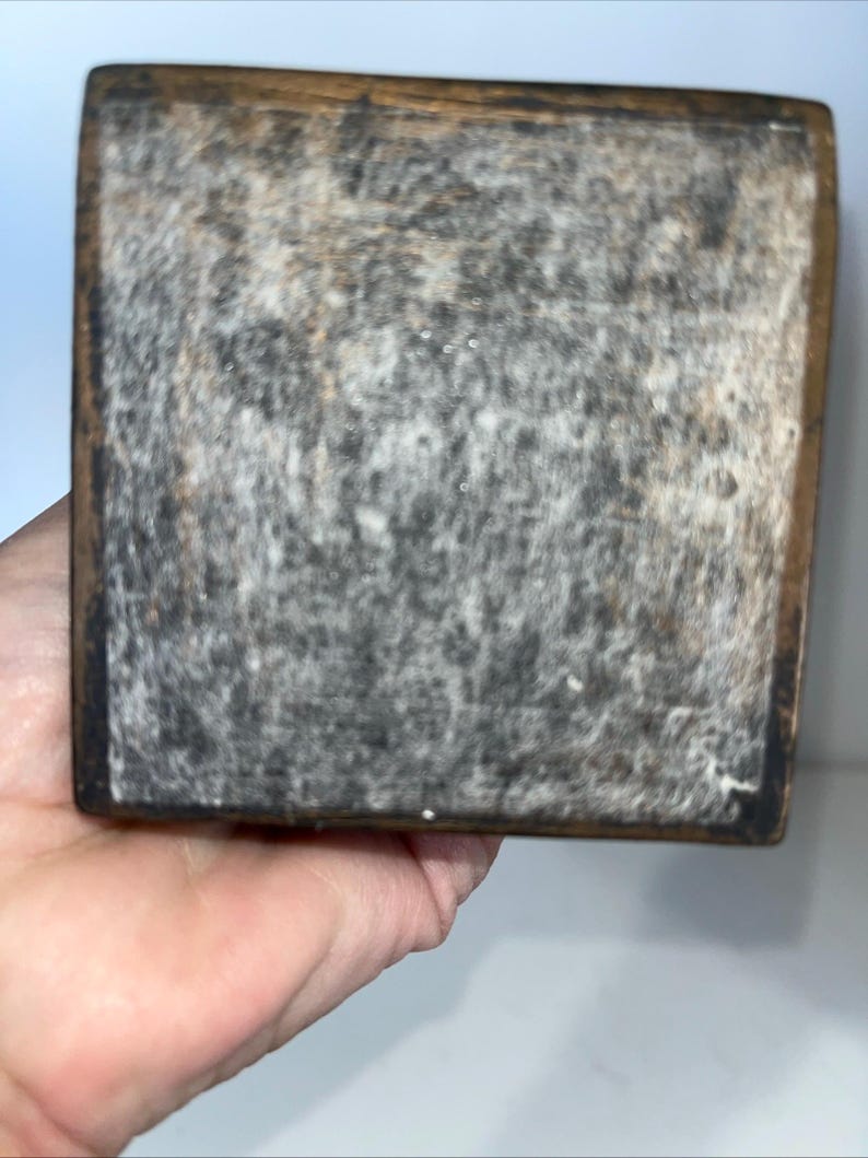 May include: Close-up of a weathered, rectangular wooden block with a dark, textured surface. The edges are framed with a darker wood. The block is held in a hand.