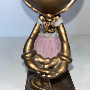 May include: A bronze-coloured figurine of a character with a large head and outstretched arms. The figure wears a pink tutu and a white and gold bow. The base is rectangular and has the Disney logo.
