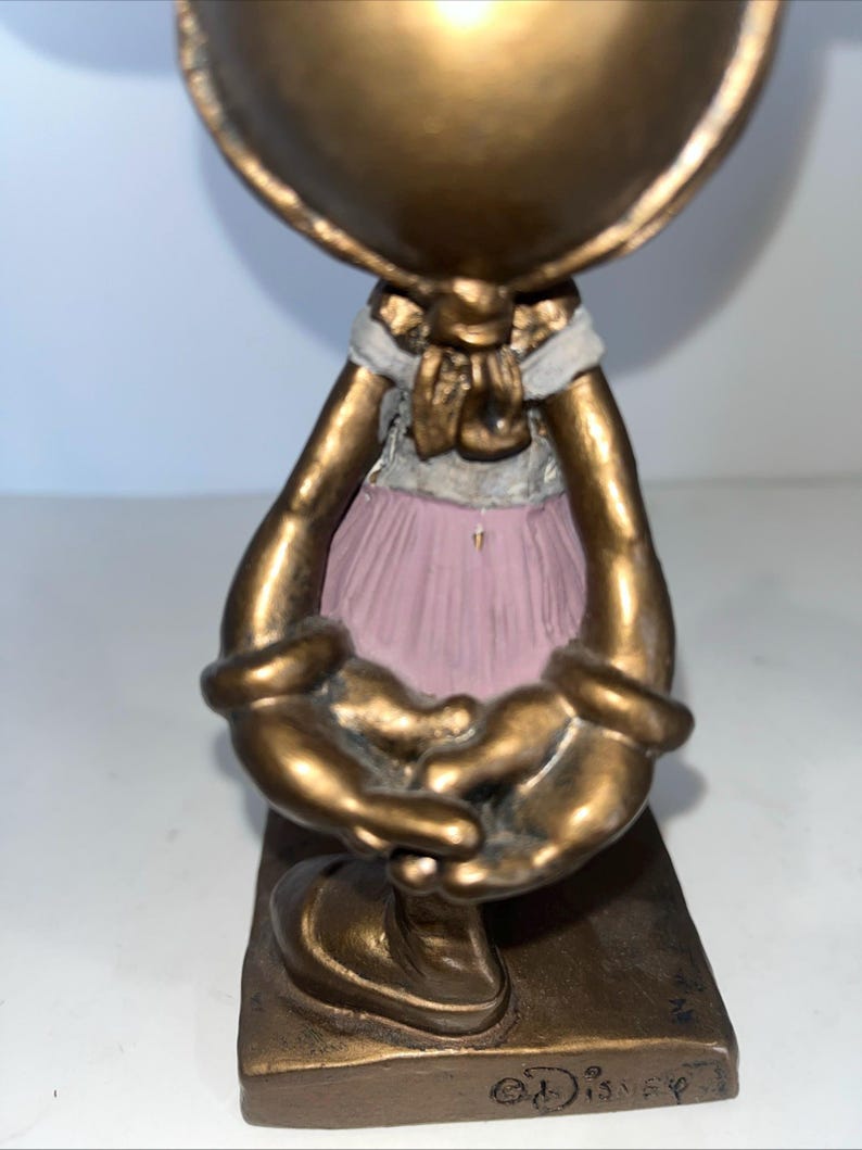 May include: A bronze-coloured figurine of a character with a large head and outstretched arms. The figure wears a pink tutu and a white and gold bow. The base is rectangular and has the Disney logo.