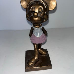 May include: A bronze-effect figurine of Minnie Mouse in a ballet outfit. The figure has large, round ears and a slightly downcast expression. She wears a white top with a silver vest and a pink tutu. The figurine stands on a square base.