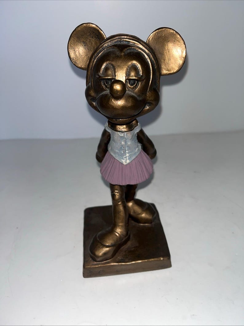 May include: A bronze-effect figurine of Minnie Mouse in a ballet outfit. The figure has large, round ears and a slightly downcast expression. She wears a white top with a silver vest and a pink tutu. The figurine stands on a square base.