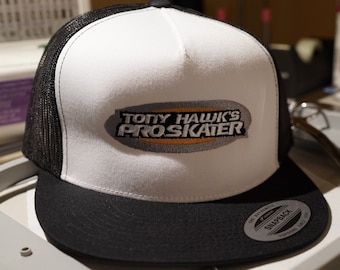 May include: A white and black trucker hat with a black mesh back. The front of the hat has a grey and orange embroidered logo that says "Tony Hawk's Pro Skater".