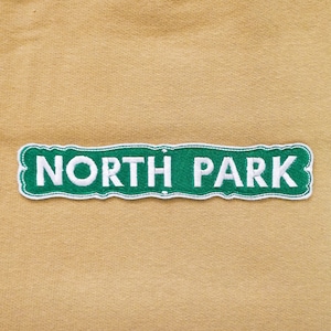 May include: A tan t-shirt with a green and white embroidered patch that says "NORTH PARK".