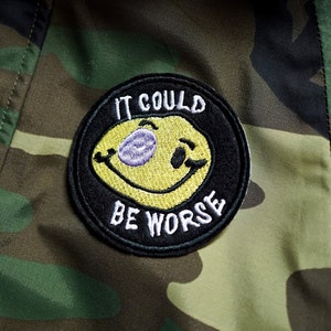 May include: A black and yellow embroidered patch with a smiley face and the text "IT COULD BE WORSE" on a camouflage background.