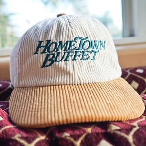 May include: A white and tan corduroy baseball cap with the text "HOMETOWN BUFFET" embroidered in green on the front.
