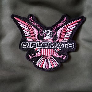 Könnte beinhalten: Ein gestickter Aufnäher in Schwarz und Pink mit einem Weißkopfseeadler mit ausgebreiteten Flügeln und dem Wort "DIPLOMATS" in weißen Buchstaben.