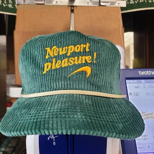 以下が含まれることがあります： 「Newport pleasure!」の文字が黄色で書かれた、緑色のコーデュロイバケットハット。つばの周りには白いロープのディテールがあり、黄色のスウッシュロゴが付いています。帽子は白と青の表面にあります。
