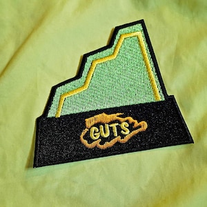 May include: A green and yellow embroidered patch with a mountain design and the word "GUTS" in a cloud shape.