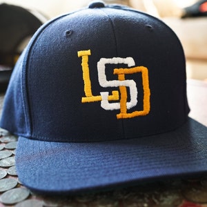 Pode incluir: Um boné de beisebol azul marinho com um logotipo "SD" bordado em branco e amarelo.