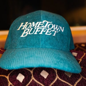 May include: A teal blue corduroy baseball cap with white embroidered text that reads "Hometown Buffet".