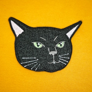 Op de afbeelding: Zwarte geborduurde kattenkop patch met groene ogen en witte snorharen.