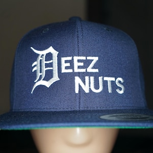 Puede incluir: Gorra de béisbol azul marino con visera plana y parte inferior verde. La gorra presenta el texto blanco "EEZ NUTS", con una letra "D" estilizada al principio. La gorra es de estilo snapback.