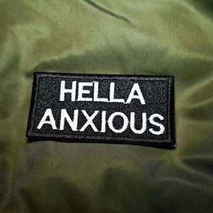 May include: A black patch with white stitching that reads "HELLA ANXIOUS" is sewn onto a green jacket.