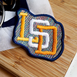 Op de afbeelding: Geborduurde patch met de letters "LS" in wit en oranje, omlijnd met een blauwe rand. De patch heeft een getextureerde, veelkleurige achtergrond. Schaar en stofresten zijn zichtbaar, wat wijst op een knutsel- of naaiproject.
