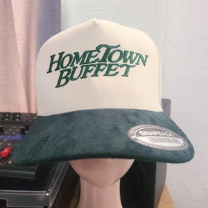 Może przedstawiać: Biało-zielona czapka z daszkiem z haftowanym napisem "Hometown Buffet" z przodu. Czapka ma zielony daszek ze skóry zamszowej z etykietą "Snapback".