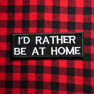 Puede incluir: Un parche negro con texto bordado en blanco que dice "I'D RATHER BE AT HOME" está centrado en una tela a cuadros roja y negra. El parche es rectangular.