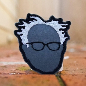 May include: A gray embroidered patch of a person with white hair and black-rimmed glasses.